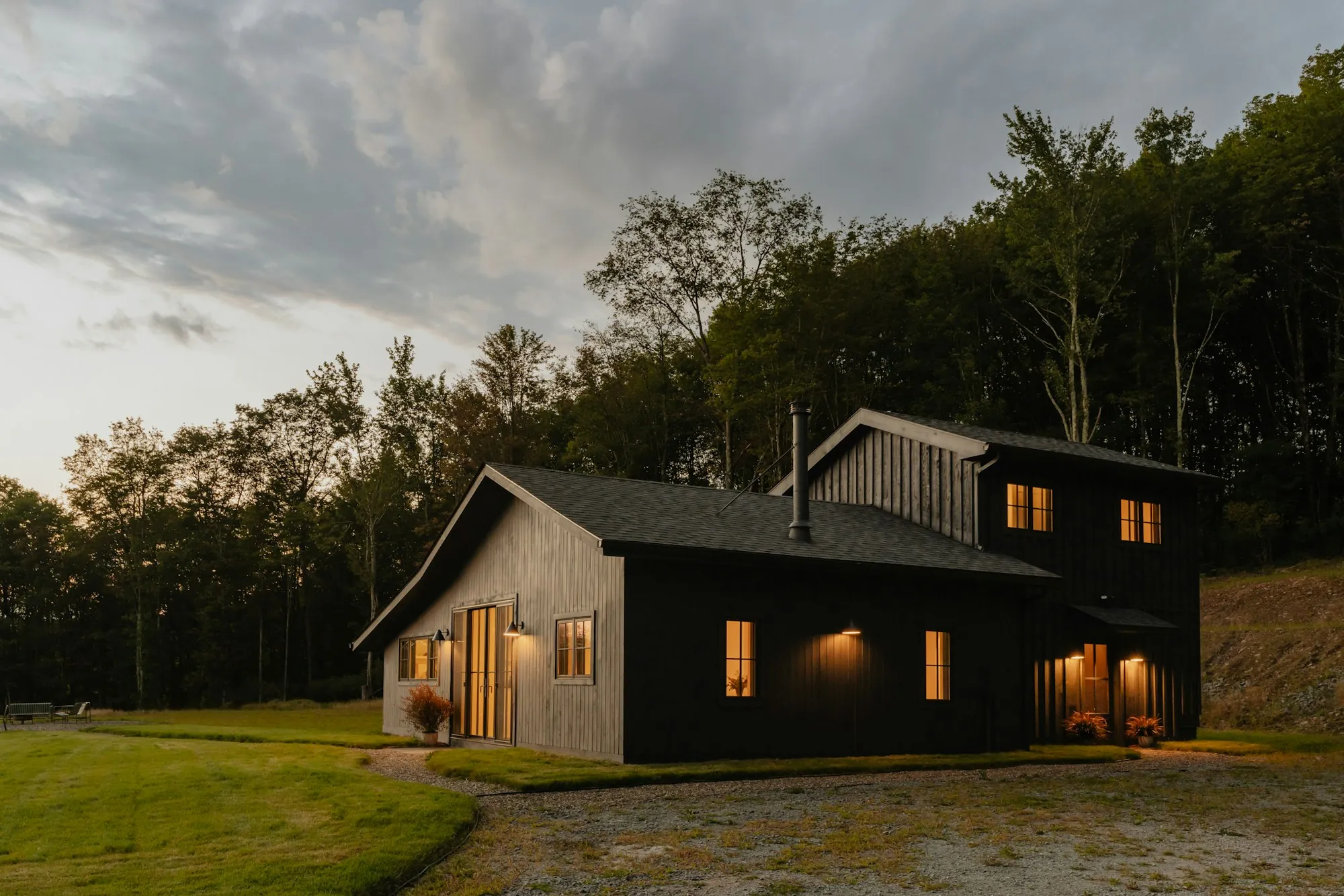 Dark-sided country home glowing at dusk beside a forest